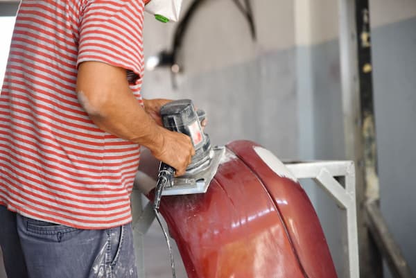 A technician feathers down a maroon vehicle panel with an orbital sander