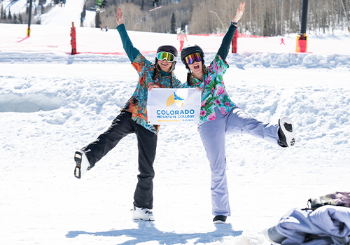 Alumni holding a CMC Adventure Flag at the base of a ski area.
