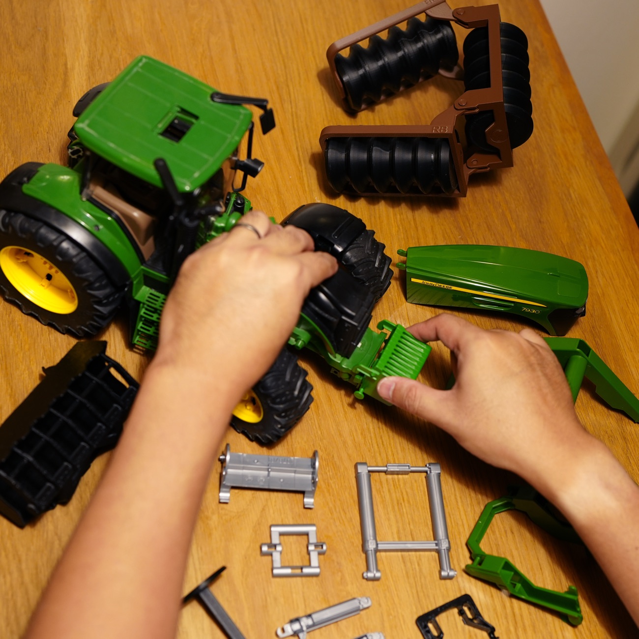 Hands reassembling a green Bruder John Deere toy tractor, with spare and 3D printed parts spread across a wooden table