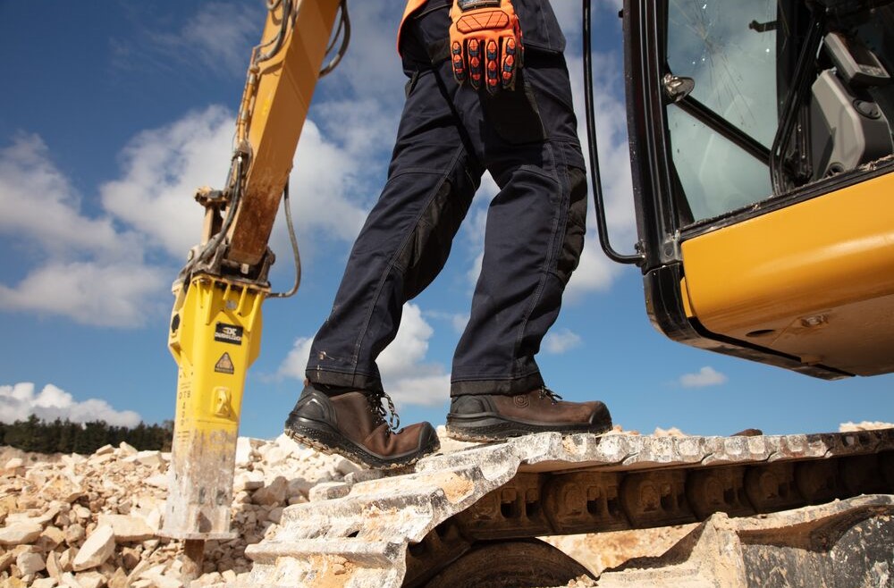 Arbeiter in Sicherheitsschuhen auf unebenem Untergrund neben Baumaschine im Außeneinsatz.